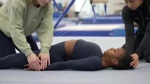 AI Video from prompt: A gymnast lies supine on the ground. Two assisti