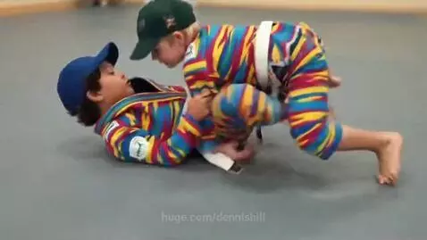 Two young boys in colorful BJJ gis and baseball caps practice ground grappling on a mat.