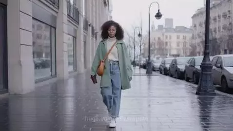 Young woman with curly hair and glasses walks down a wet city sidewalk in a green coat, holding a coffee cup and bag.