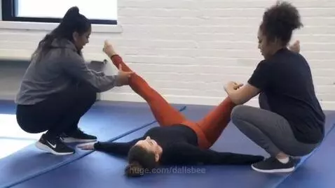 Gymnast lying on back on blue mat, assisted into a full split by two kneeling individuals.