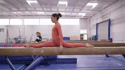 Gymnast in orange leotard doing splits on a balance beam, assisted by another gymnast.