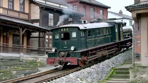 Dark green vintage steam train with gold accents ascending a stone-lined track between historical buildings.