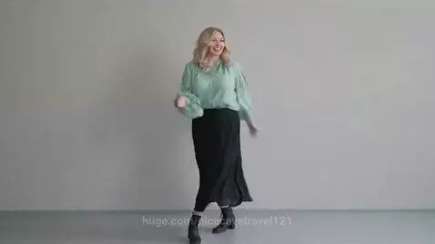 Blonde woman with wavy hair in a green blouse and black skirt dancing joyfully in a minimalist room.