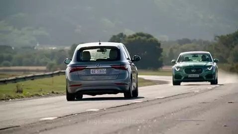 A grey Yugo hatchback drives away from the camera on a rural road, while a green 1990 Jaguar XJ sedan approaches.