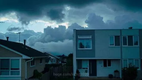 Time-lapse of dramatic clouds over houses, zooming into a window with human silhouettes.
