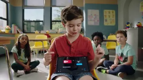 An 8-year-old boy in a colorful classroom enthusiastically explains AI using a tablet to his friends.