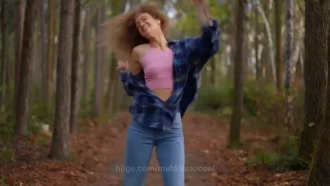 Young woman with long, curly, light brown hair dancing in a forest wearing a pink crop top and blue jeans.