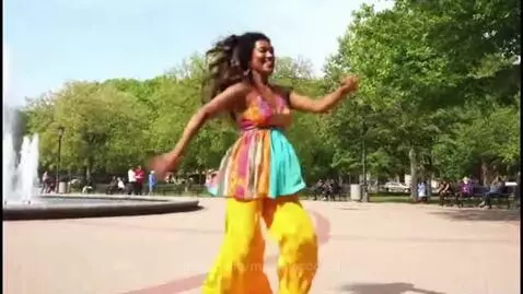 Woman with dark curly hair dancing energetically in a park, wearing a colorful top and yellow pants, with a fountain in the background.