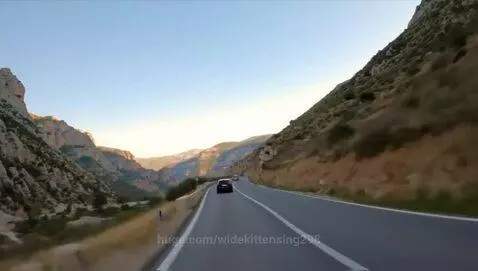 First-person view from a car driving on a paved mountain road with steep, rocky hills on either side.