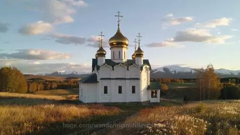 A white Russian Orthodox church with golden domes stands in a golden autumn field, with mountains in the background.