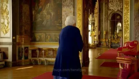 Elderly woman in a regal pose gazes out a window in a richly decorated baroque palace interior with warm lighting.