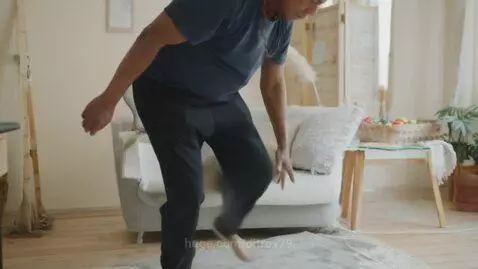 Man with dark curly hair in a blue shirt and black pants stretching his leg on a rug in a living room.