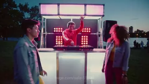 DJ with headphones and red jacket playing 80s music at a mobile disco in a park with people dancing.