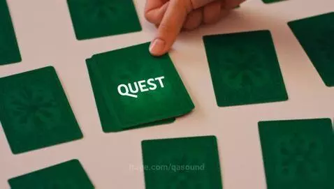 Top-down view of a hand picking up a green card with glowing 'QUEST' text inside.