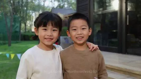 A young Asian boy and girl stand side-by-side outdoors, smiling at the camera, with a grassy yard and house in the background.