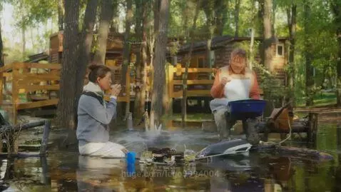 Couple sitting in shallow forest water, woman drinks from cup, man holds container, wooden cabins visible.