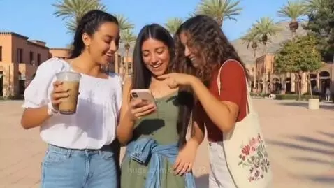 Three young Hispanic women standing outdoors, laughing together while looking at a smartphone.