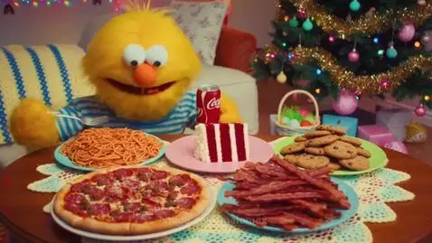 Yellow puppet with blue striped shirt eating spaghetti at a table with pizza, cake, bacon, and cookies, with Christmas and Easter decorations.