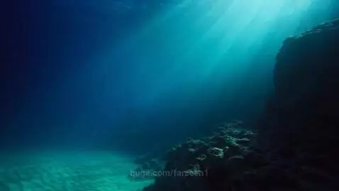 Sunlight rays shining through deep blue ocean water, illuminating a sandy seabed and dark rock formations.