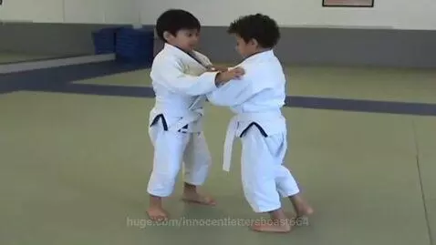 Two young boys in white BJJ gis and cropped pants practice grappling techniques in a dojo.