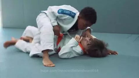 Two young boys in white BJJ gis grappling on a blue mat, one in a dominant position.