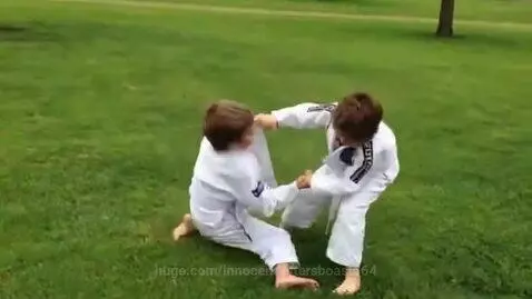 Two young boys in white BJJ gis grappling on a green grass field.