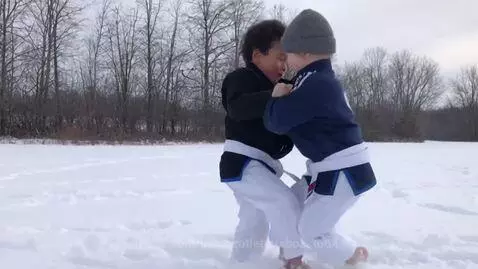 Two young boys wearing BJJ gis and white belts grapple barefoot in a snowy field with bare trees.