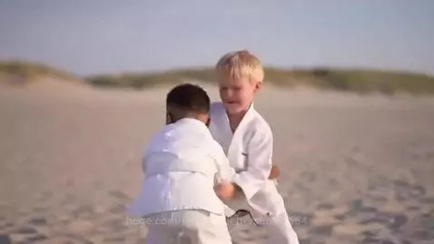 Two young boys in white BJJ uniforms grappling on a sandy beach with dunes in the background.