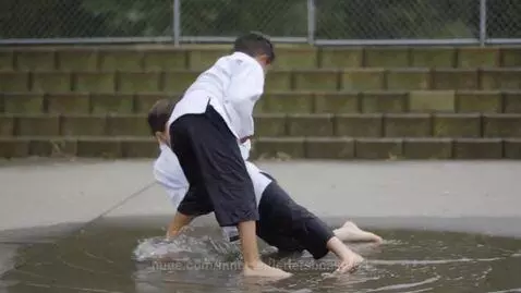 Two young boys in white BJJ jackets and black pants grappling in a shallow puddle, splashing water.
