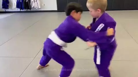 Two young boys in purple BJJ gis with white belts grappling on a mat, one performing a sweep.