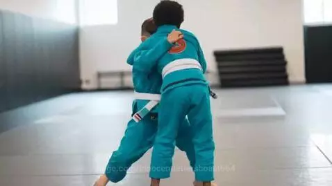 Two young boys in turquoise BJJ uniforms with white belts grappling on a mat during a training session.