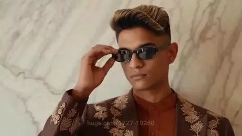 Young man with stylish haircut, sunglasses, and embroidered brown jacket posing against a marbled wall.
