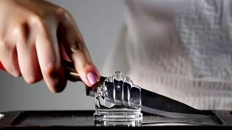 Close-up of a hand holding a knife cutting a transparent glass chess queen on a black cutting board.