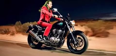 Man with green hair dressed as Joker riding a black Harley-Davidson motorcycle on a desert road at night.
