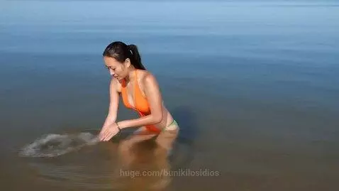 Woman in orange swimsuit kneeling in shallow seashore water, splashing and creating ripples.