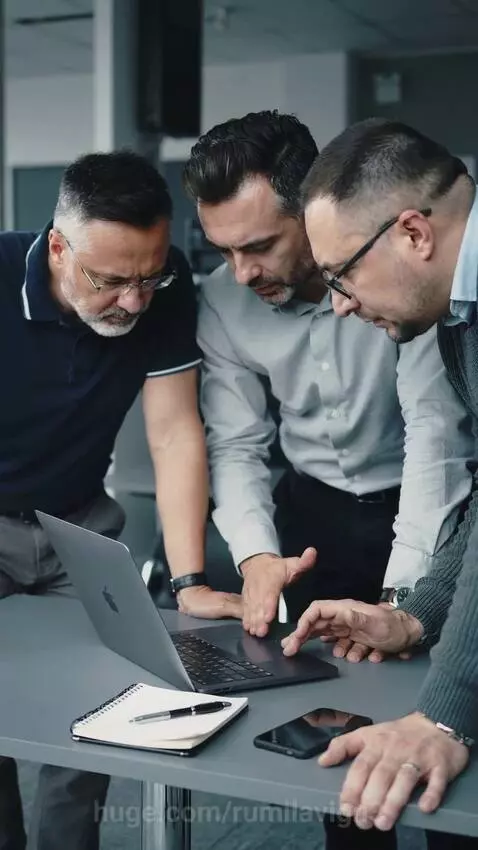 Three men gathered around a silver MacBook on a grey table, pointing and touching the trackpad, engaged in discussion.