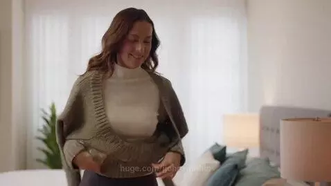 Woman with long brown hair putting on a chunky, olive-green V-neck sweater over a white top in a bright bedroom.