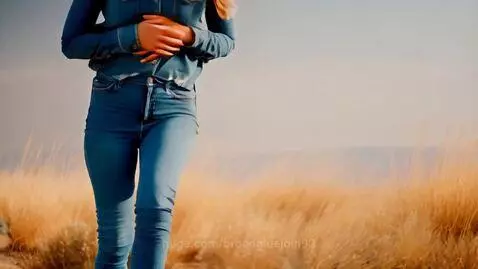 Woman in denim shirt, jeans, and cowgirl boots walking in a dry, grassy field.