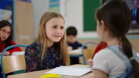 A young girl in a classroom throws a yellow object, causing white fluffy material to explode around her head.