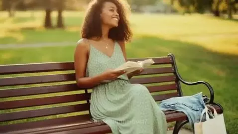 Young woman with curly hair reading a book on a wooden park bench in a sunny park.