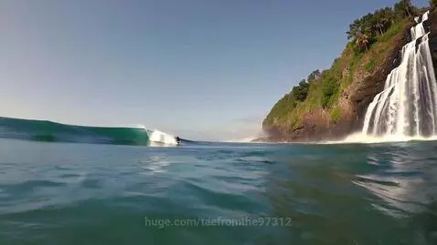 Surfer in black wetsuit riding a wave near a multi-tiered waterfall cascading into the ocean.