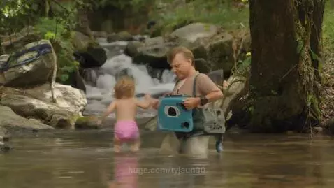 Toddler in diaper walking in a shallow forest stream towards a man holding a backpack.