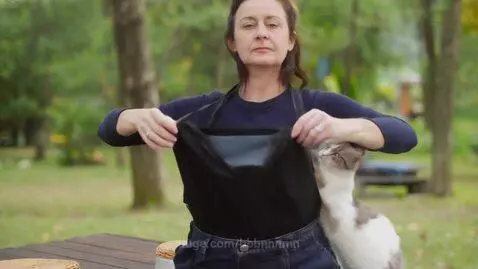 Woman in blue shirt and jeans outdoors in a park, placing a white and grey cat into a fabric carrier worn around her waist.