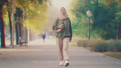 Woman wearing a hijab and green sweatshirt walks in a park, holding a coffee cup and smiling.
