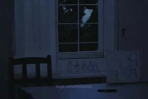 A child looks out a window at dusk, with drawings of a family on a table, one figure faded.