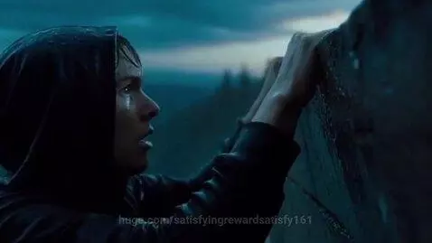 Side view of a young man in a black hoodie clinging to a wet ledge, looking up, in the rain.