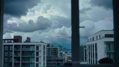 Silhouettes of people visible behind an apartment window with a cloudy sky and multi-storey buildings in the background.