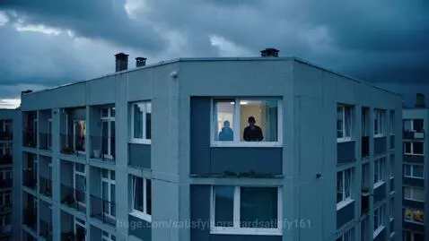 Silhouettes of two people visible through a brightly lit apartment window, looking out at a cloudy sky.