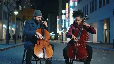 Two musicians playing cellos on a city street at night, with illuminated buildings in the background.