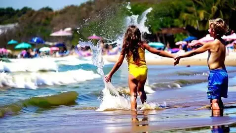 Two children, a girl in yellow and a boy in blue, play happily in the shallow water at a sunny beach.